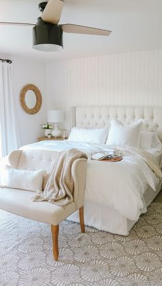 a white bed sitting next to a window in a bedroom under a ceiling fan on top of a wooden table