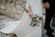 a bride and groom holding hands while standing next to each other in front of a stone wall