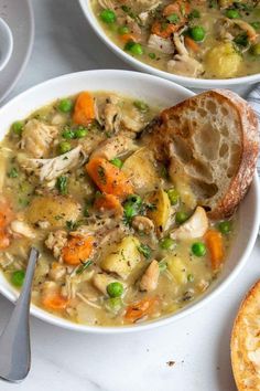 two bowls of chicken and vegetable soup on a white table with bread, spoons and napkin