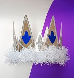 three paper crowns with blue hearts on them sitting on a purple and white wall next to some feathers