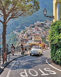 a car driving down a street next to a lush green hillside covered in trees and buildings