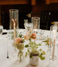 a table topped with vases filled with flowers and greenery next to candles on top of a white table cloth