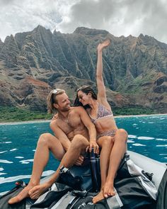 a man and woman sitting on the back of a boat in the ocean with mountains behind them