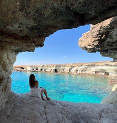 a woman sitting on the edge of a cave looking out at the blue water and cliffs