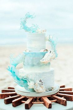 a three tiered wedding cake with blue and white frosting on the top is surrounded by seashells