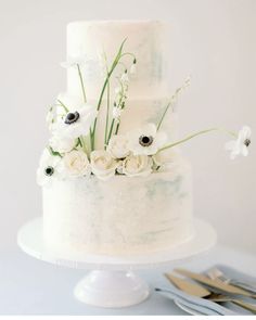 a wedding cake with white flowers and greenery on the top is surrounded by silverware