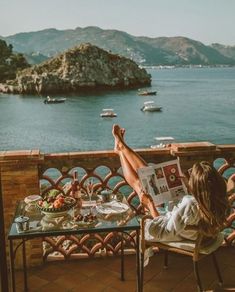 a woman sitting at a table reading a magazine overlooking the ocean and boats in the water