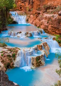 blue water flowing down the side of a cliff in front of trees and rocks, surrounded by red rock formations