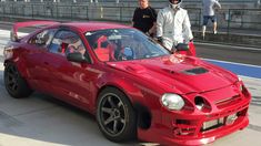 two men standing next to a red sports car on a race track with people looking at it