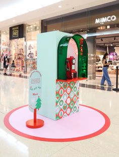 a green and red machine in the middle of a shopping mall with people walking by