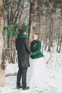 a man and woman standing next to each other in the snow holding hands with trees behind them