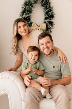 a man, woman and child sitting on a white chair in front of a christmas wreath