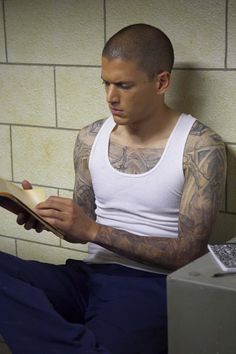 a man with tattoos sitting on a toilet reading a book while looking at his phone