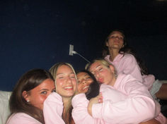 four girls in pink sweatshirts hugging each other on a bed with their arms around one another