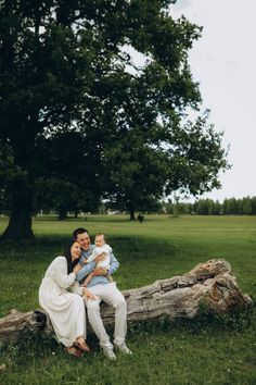 a man and woman holding a baby sitting on a log in the grass near a tree