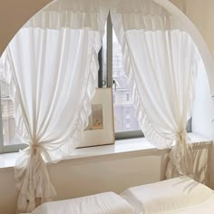 two white beds sitting under a window with drapes on the sides and curtains hanging from them