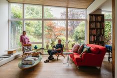 two men are sitting in the living room with bookshelves and large glass windows