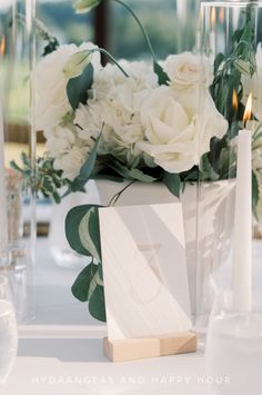 white flowers and greenery sit in a vase on a table with candles, napkins and cards