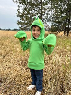 a young boy in a green frog costume standing in tall grass with his hands up