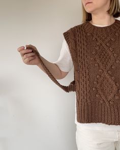 a woman wearing a brown knitted sweater and holding something in her hand while standing against a white wall