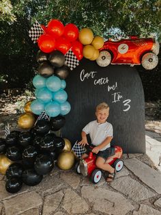 a young boy sitting in front of a chalkboard with balloons and race cars on it