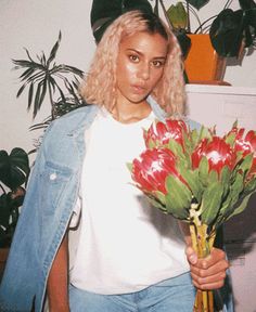 a woman holding a bouquet of flowers in her hand and looking at the camera while standing next to a potted plant