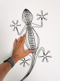 a hand is pointing at a wire lizard on a white surface with black and silver lines