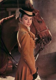 a woman standing next to a brown horse wearing a yellow dress and black hat with her hand on the bridle