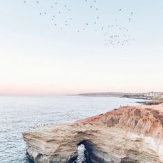 birds are flying over the water near an arch in the rock formation on the beach