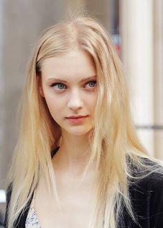 a woman with long blonde hair and blue eyes looks at the camera while standing in front of a building