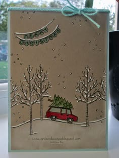 a handmade christmas card with a red car carrying a tree in the snow, on a window sill