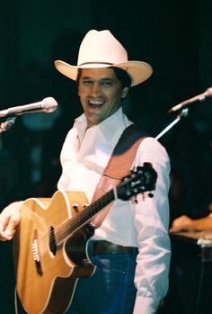 a man wearing a cowboy hat and holding a guitar in front of microphones on stage