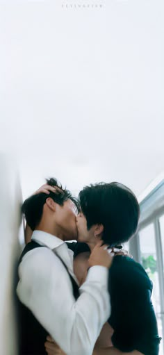 two people are kissing each other in front of a white wall with windows behind them