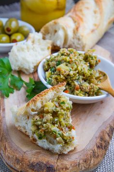 a wooden cutting board topped with bread and bowls filled with different types of food next to olives