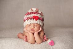 a newborn baby wearing a knitted hat with hearts on it's side and her hands under her chin