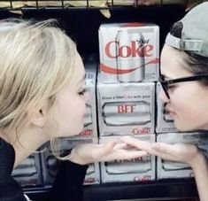 two young women are looking at each other in front of soda coolers with boxes behind them