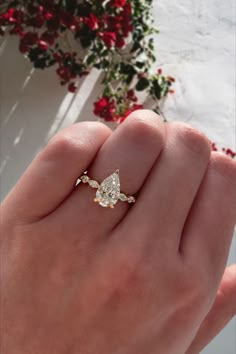 a woman's hand with a diamond ring on top of her finger and red flowers in the background