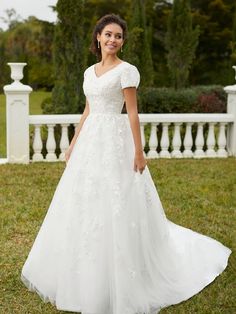 a woman in a white wedding dress standing on the grass with her hands in her pockets