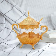 a golden tea pot and saucer sitting on top of a bed next to an open book