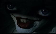 an evil looking clown with blue eyes and white teeth in the dark, staring at the camera