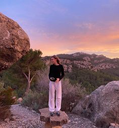 a woman standing on top of a large rock next to a tree filled hillside at sunset