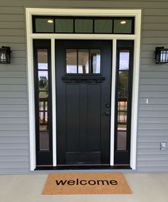 a welcome mat is on the front door of a gray house with black doors and windows