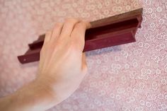 a hand holding a piece of wood on top of a pink wallpapered floor