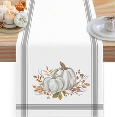 a table runner with white pumpkins and leaves on it, next to a candle holder