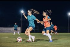 two girls are playing soccer at night on the field with lights in their hands and one girl is running after the ball