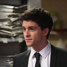 a young man wearing a suit and tie in front of stacks of papers on a desk