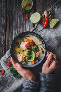 a person holding a bowl of soup on top of a wooden table with limes