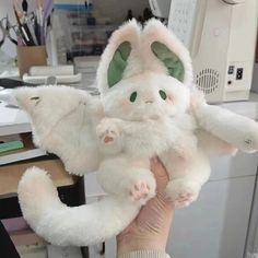 a hand holding a white stuffed animal with green eyes and tail sitting on top of a desk