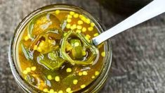 a jar filled with pickles and corn on top of a wooden table next to a spoon