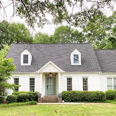 a white house with a gray roof and black shutters on the front door is surrounded by lush green trees